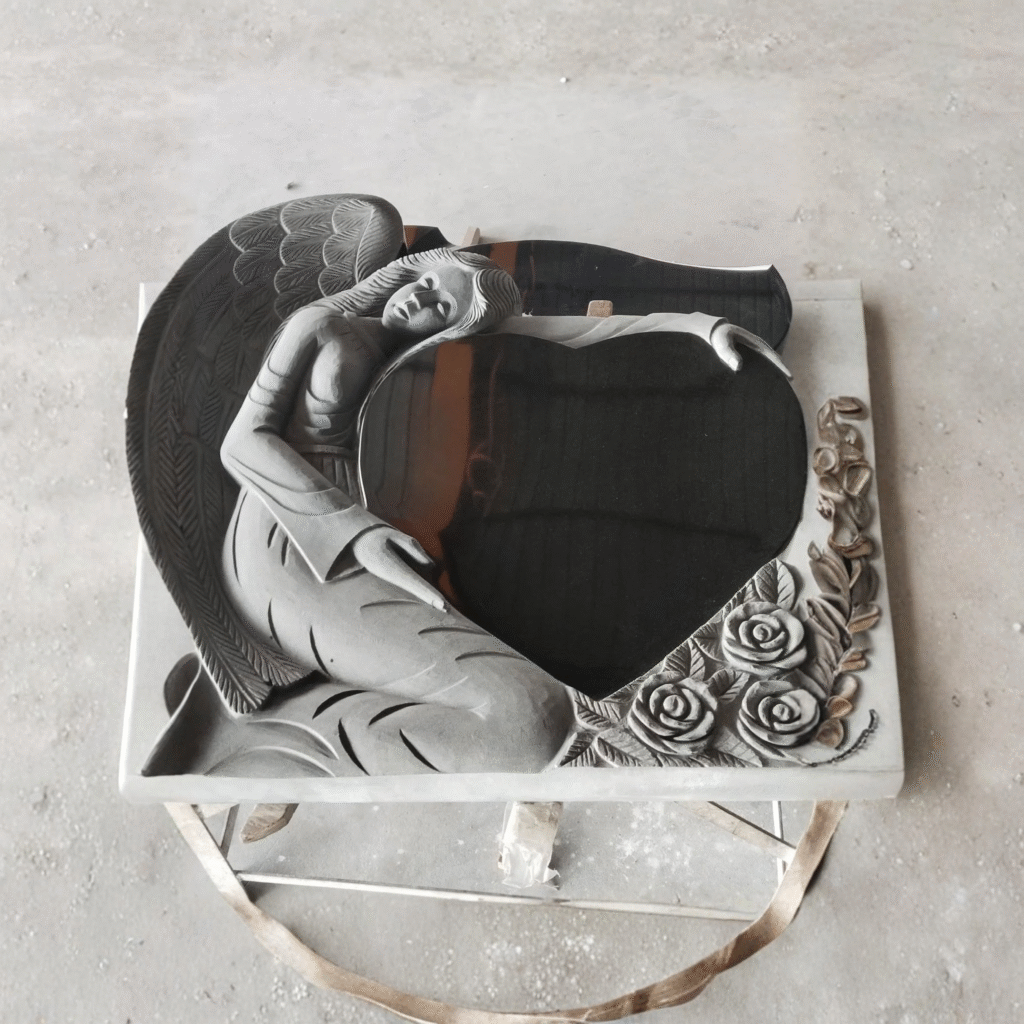 High-polished black granite headstone featuring a masterfully hand-sculpted kneeling angel embracing a polished heart, accented with detailed carved roses at the base