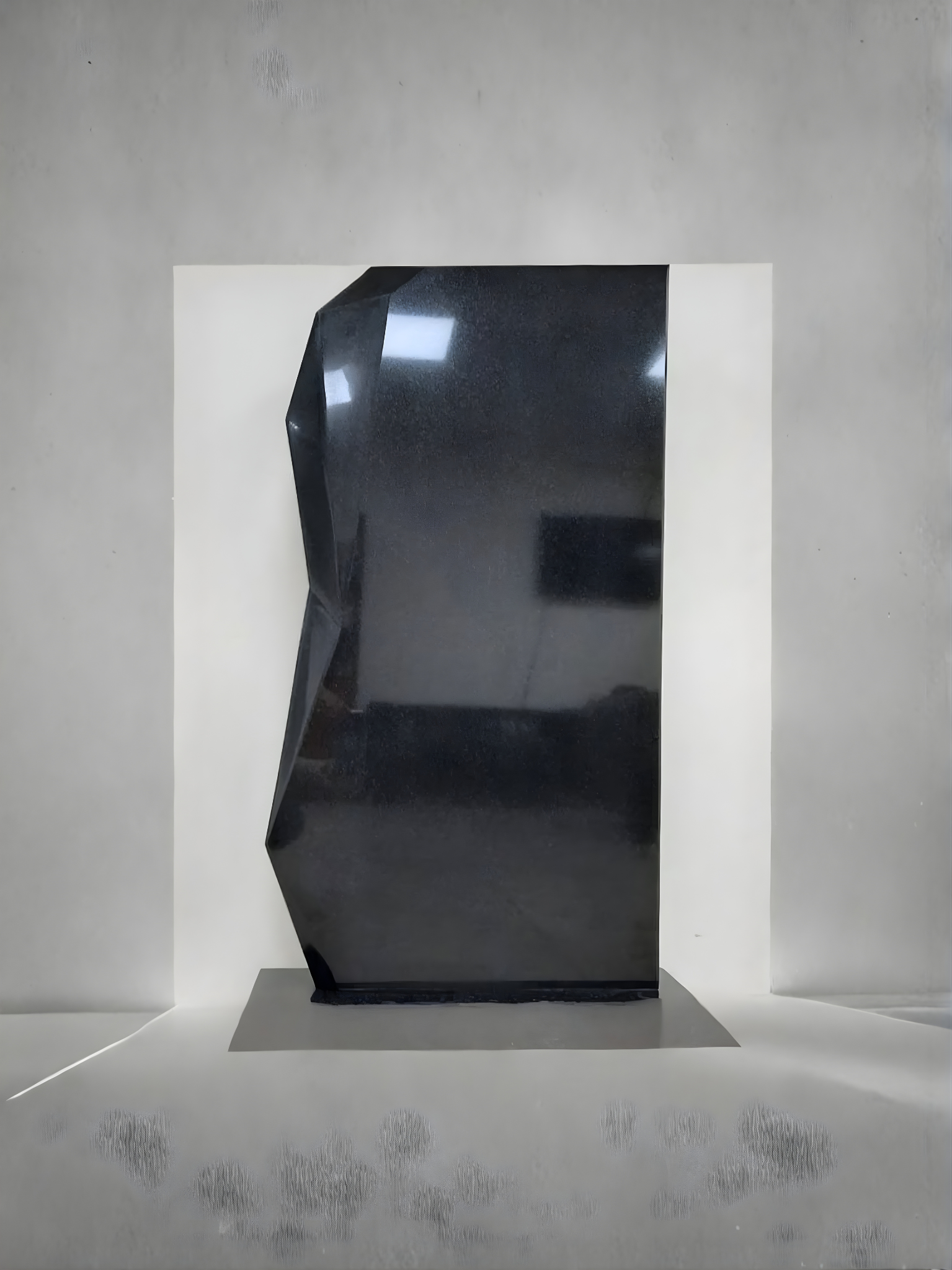 A modern geometric headstone made of polished black granite with fine speckles, featuring complex faceted planes on one side and a smooth flat surface on the other, resting on a matching base.